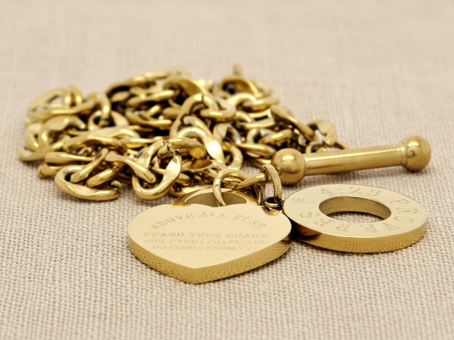 Gold chain with heart-shaped tag and round disc on a beige fabric background